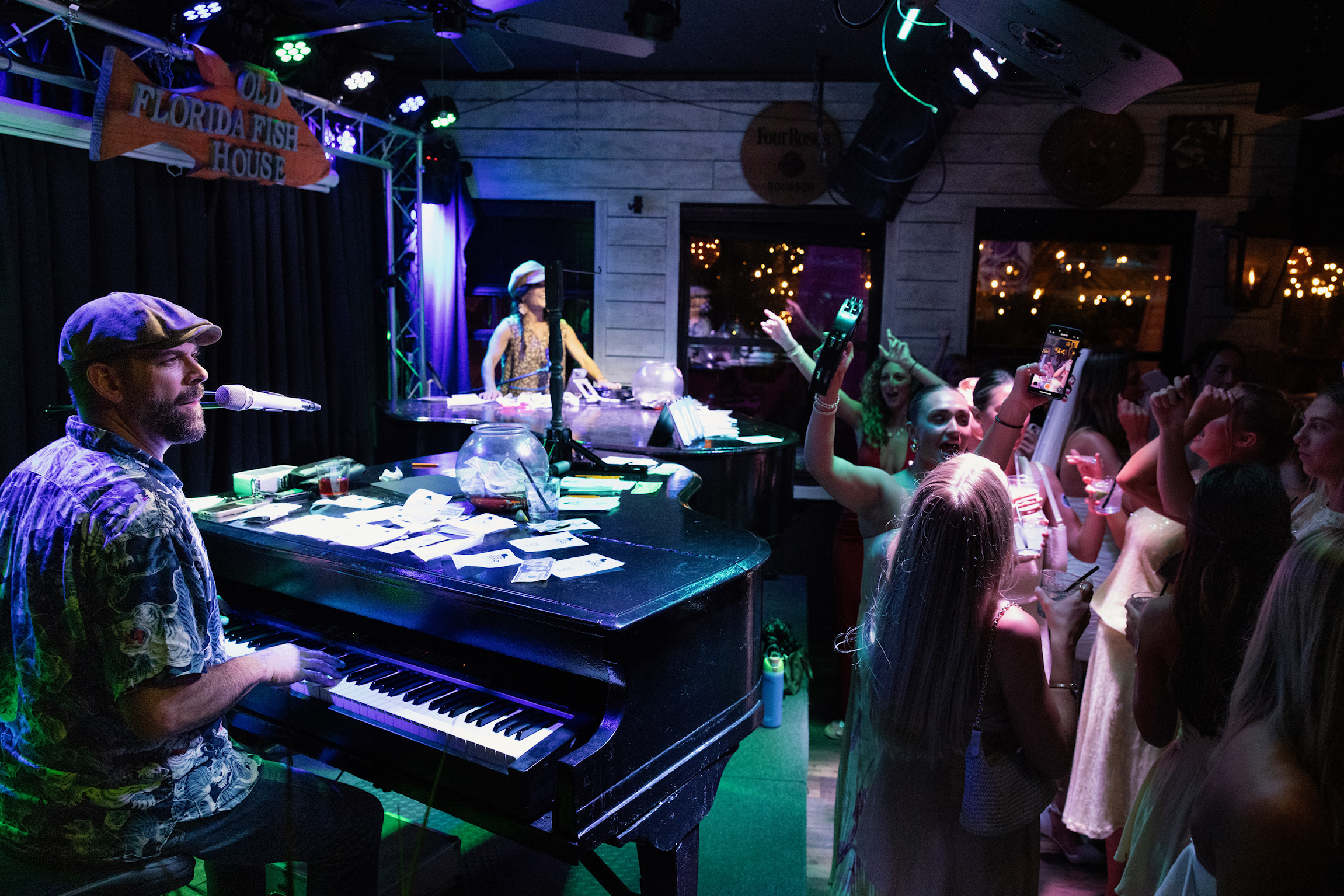 Guests gather around the dueling pianos stage at Old Florida Fish House as musicians perform live, with audience members singing along, raising drinks, and capturing the lively entertainment on their phones.